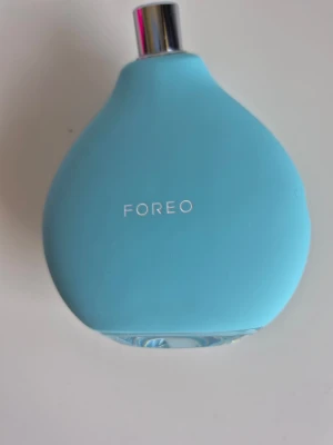 Blå FOREO ansiktsapparat - Säljer en blå ansiktsapparat från FOREO med rundad, droppformad design. Tillverkad i silikon med metallisk topp och medföljande vit laddningssladd. Perfekt för hudvårdsrutinen och enkel att använda. Original pris 1 595kr oanvänd 