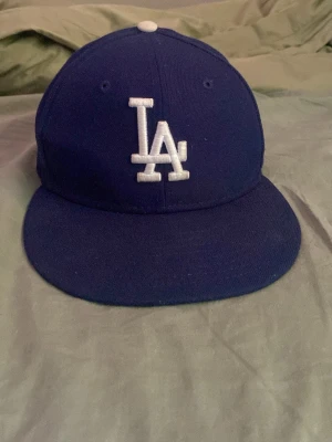 Blå LA keps - Snygg blå keps med LA Dodgers-logga broderad i vitt framtill och MLB-logga baktill. Klassisk rak skärm och ventilerande hål upptill. Perfekt för dig som gillar sportig streetstyle och vill representera Los Angeles Dodgers.