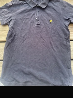 Mörkblå pikétröja från Lyle & Scott - Klassisk mörkblå pikétröja från Lyle & Scott med gul logga på bröstet. Tröjan har krage, knappar framtill och är kortärmad. Perfekt till jeans eller shorts för en clean och enkel stil.