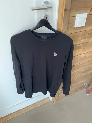Svart långärmad tröja med tryck - En svart långärmad tröja i mjukt material med rund halsringning och ett litet tryckt motiv på bröstet. Enkel och stilren design som passar till det mesta. Perfekt basplagg för garderoben.