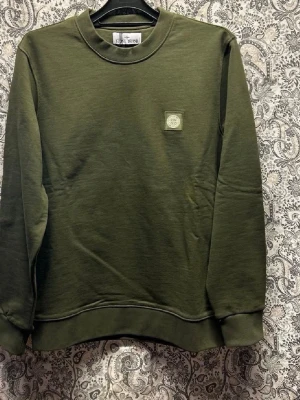 Olivgrön sweatshirt med logopatch - Olivgrön sweatshirt från Stone Island med rund hals och ribbade muddar. Tröjan har en diskret logopatch på bröstet och är tillverkad i mjukt bomullsmaterial. Perfekt för en avslappnad och stilren look.