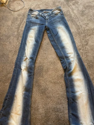 Blå bootcut jeans med blekta detaljer - Säljer ett par blå bootcut jeans med tydliga blekta partier längs benen. Jeansen har klassisk femficksdesign och låg midja. Materialet är denim och passformen är utsvängd nertill för en retro vibe.