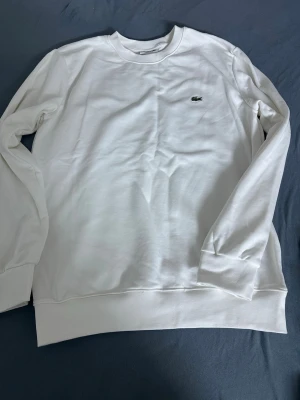 Vit sweatshirt från Lacoste - Klassisk vit sweatshirt från Lacoste med den ikoniska gröna krokodilloggan broderad på bröstet. Tröjan har rund halsringning, långa ärmar och ribbade muddar. Perfekt för en clean och stilren look.