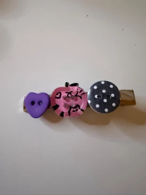 Handgjord hårklämma med knappar💜🖤🩷 - Unik hårklämma dekorerad med tre stora knappar: en lila hjärtformad, en rosa med svart mönster och en svart med vita prickar. Klämman är i metall och ger en lekfull och kreativ touch till din frisyr.