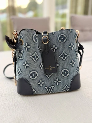 Snygg, trendig, modern och klassisk denim handväska med fina detaljer - Snygg handväska som är lika trendig som den är klassisk i blått denimtyg med ikoniskt monogrammönster och mörkblå läderdetaljer. Väskan har guldfärgade metalldetaljer och ett handtag med svartvitt mönster. Perfekt accessoar för dig som vill sticka ut. 