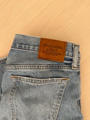 Ljusblå jeans från Abercrombie & Fitch - Säljer ett par klassiska ljusblå jeans från Abercrombie & Fitch. Jeansen har en rak passform, fem fickor och snygga kontrastsömmar. Tillverkade i slitstark denim med en vintagekänsla. Perfekta för en avslappnad och trendig look.