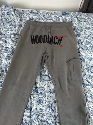 Gråa mjukisbyxor från Hoodrich - Gråa mjukisbyxor från Hoodrich med stor svart och röd logga framtill. Byxorna har dragsko i midjan, sidofickor och en extra ficka med dragkedja på benet. Tillverkade i mjukt och skönt material, perfekta för en chill look.Tillsammans med en hoodie som är väldigt skön och stor text där fram och en ficka på magen med dragkedja 