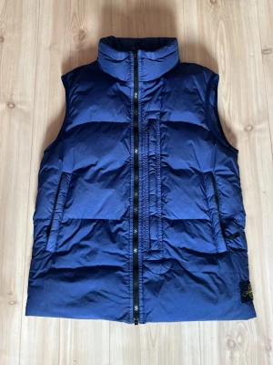 Blå dunväst från Stone Island - Snygg blå dunväst från Stone Island med hög krage och dragkedja framtill. Västen har två stora fickor och klassisk logotyp på vänster sida. Perfekt för lager-på-lager och streetwear-stil.