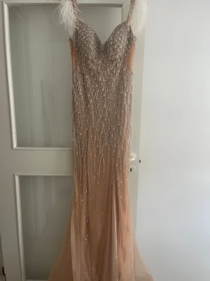 Glittrig långklänning med fjädrar - Säljer en elegant långklänning i beige med glittriga paljetter och stenar över hela klänningen. Axelbanden är dekorerade med vita fjädrar. Klänningen har en figurnära passform och djup v-ringning. Perfekt för ba, bröllop eller fest. Använd endast en gång. Pris är disskuterbart. går att testa och köpa på plats. Skicka samma dag. Bekväm och inte tung klänning. Köpt i klänningbutiken Sasha i Södertälje