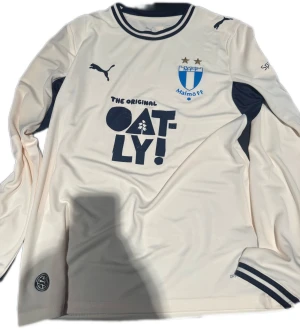 Malmö FF bortatröja långärmad Puma - Långärmad bortatröja från Malmö FF i ljus beige färg med mörkblå detaljer. Tröjan har klubbmärke, Puma-logga och Oatly-tryck på bröstet. Materialet är lätt och andas, perfekt för fotboll och träning. Snygga ribbade muddar och kontrastpaneler under armarna.