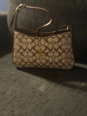 Beige och brun axelväska Coach - Snygg axelväska från Coach i beige och brunt med det klassiska C-mönstret över hela väskan. Väskan har guldfärgade detaljer och justerbart axelband. Tillverkad i canvas med läderdetaljer. Perfekt storlek för det viktigaste och ger en trendig look.