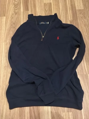 Mörkblå halvzip tröja från Polo Ralph Lauren - Mörkblå långärmad tröja med halv dragkedja från Polo Ralph Lauren. Tröjan har en klassisk röd broderad logga på bröstet och ribbade muddar vid ärmslut och nederkant. Perfekt för en avslappnad och stilren look.