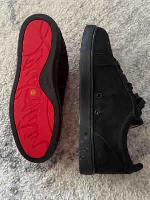 Svarta loubs med röd sula - Stilrena svarta sneakers med klassisk låg profil och snörning. Skorna har en ikonisk röd sula och är tillverkade i mocka och läder. Perfekta för dig som gillar en clean och modern look med en twist. står 40 men den passar 41.