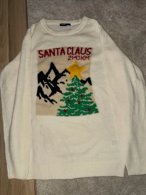 Vit jultröja med julgran och text - Vit stickad tröja med motiv av en julgran, stjärna och berg samt texten 'Santa Claus 2143 KM' på bröstet. Tröjan har rund hals och långa ärmar. Perfekt för julens alla tillfällen och mysiga vinterdagar. Priset kan diskuteras och sänkas till en viss summa.