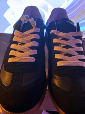 Svarta sneakers med vita snören - Snygga svarta sneakers med vita snören och detaljer i mocka och läderimitation. Yttersulan är i gummi och skon har en klassisk låg siluett som passar till många olika stilar. Perfekta för dig som vill ha en clean och tidlös look.