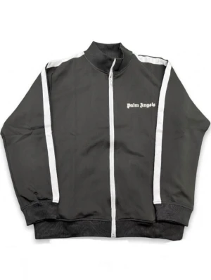 Svart track jacket från Palm Angels - Snygg svart track jacket från Palm Angels med vita ränder längs ärmarna och vit dragkedja. Tröjan har hög krage, ribbade muddar och logga på bröstet. Perfekt för en sportig och trendig look.