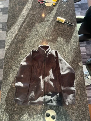 Mörkgrå fleecejacka med dragkedja - Mysig mörkgrå fleecejacka med hög krage och hel dragkedja framtill. Jackan har två sidofickor med dragkedja och en enkel, stilren design. Perfekt för kyliga dagar och enkel att matcha med olika outfits.
