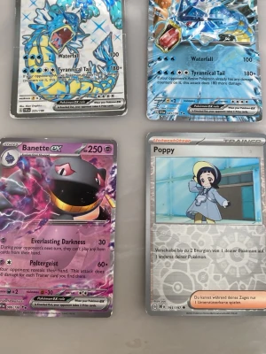 Fyra Pokémonkort EX och Trainer - Säljer fyra Pokémonkort: två Gyarados EX med blått holografiskt motiv, ett Banette EX med lila och svart design samt ett Trainer-kort 'Poppy' med en tecknad karaktär. Alla kort har färgstarka illustrationer och är tryckta på glansigt papp.