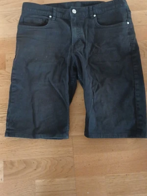 Svarta shorts med fem fickor - Säljer ett par svarta shorts med klassisk femficksdesign och knappgylf. De har en rak passform och är tillverkade i ett mjukt bomullstyg som känns bekvämt. Perfekta för dig som gillar enkel och stilren look.