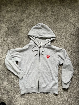 Grå hoodie Comme des Garçons Play - Säljer en grå zip-hoodie från Comme des Garçons Play med huva och fickor fram. På bröstet finns det klassiska röda hjärtat och på ryggen flera röda hjärtan samt PLAY-tryck. Tillverkad i mjuk bomull, perfekt för en chill och stilren look.