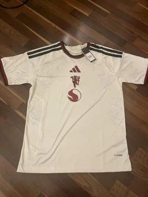 Vit Manchester United tröja Adidas - Snygg vit Manchester United tröja från Adidas med vinröda detaljer och klubbmärke på bröstet. Tröjan har svarta ränder på axlarna, rund halsringning och CLIMACOOL-material som andas. Perfekt för fotboll eller träning.