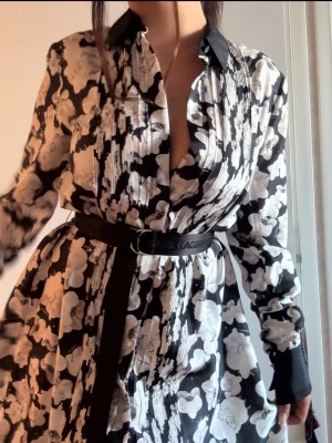 Karl Lagerfeld  - Elegant skjortklänning från Karl Lagerfeld i ett vackert monokromt blommönster. Klänningen har sofistikerade detaljer som kontrastmarkeringar i svart vid krage och manschetter, samt fina plisseringar framtill. Silhuetten markeras snyggt med det tillhörande logobältet i midjan. Ett tidlöst och lyxigt plagg som passar till både vardag och fest!  Helt ny med prislapp nypris 3995