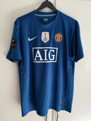 Manchester United 08/09 Away Shirt - Säljer en blå Manchester United matchtröja från Nike med AIG som sponsortryck. Tröjan har FIFA World Champions 2008-märke på bröstet, Champions League-märke på ärmen och klubbmärke på vänster sida. Perfekt för fotbollsfans och samlare.