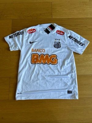 Vit Santos FC Neymar Jr matchtröja - Säljer en vit Santos FC matchtröja från Nike med Neymar Jr och nummer 11 på ryggen. Tröjan har flera sponsorloggor i svart och orange, klubbmärke på bröstet och korta ärmar. Perfekt för dig som älskar brasiliansk fotboll och vill sticka ut på planen.