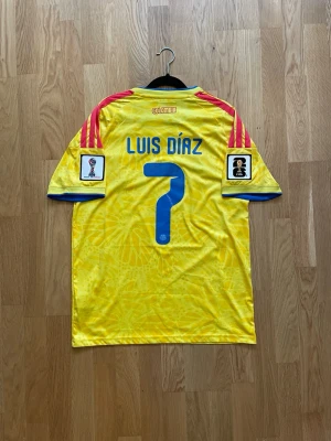 Luis Díaz #7 - M - Gul Colombia fotbollströja från Adidas med blå detaljer och röda ränder på axlarna. Nummer 7 och namnet Luis Díaz på ryggen. Världsmästerskapsmärken på ärmarna och landslagets emblem på bröstet. Tillverkad i lätt och ventilerande material.