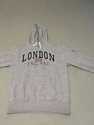 Grå hoodie med London-tryck - Säljer en grå hoodie med huva och dragsnören. På bröstet står det 'LONDON ENGLAND' i stora bokstäver och en liten brittisk flagga. Hoodien har en klassisk känguruficka och ribbade muddar. Perfekt för dig som gillar streetwear och vill ha en chill look.