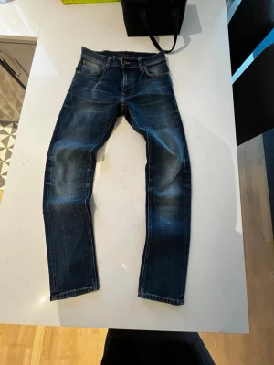 Blå jeans från Nudie Jeans - Säljer ett par blå jeans från Nudie Jeans med snyggt slitna detaljer och klassisk femficksdesign. Jeansen har raka ben och kontrastsömmar i orange. Perfekta för dig som gillar en cool och avslappnad stil. Jeansen är nersydda ungefär 5 cm passar 165-170 ungefär ser helt vanliga ut efter!