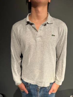 Ljusgrå långärmad piké Lacoste - Klassisk ljusgrå långärmad pikétröja från Lacoste med den ikoniska krokodilloggan på bröstet. Skjortan har krage, knappar i halsen och är tillverkad i mjuk bomull. Perfekt till jeans för en avslappnad men stilren look. Modellen är 175cm och väger70kg