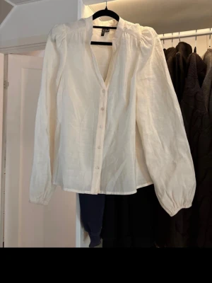 Cremevit blus från Vero Moda med puffärm - Stilren Cremevit blus från Vero Moda med knappar framtill och v-ringning. Blusen har långa puffärmar och är tillverkad i ett lätt, vävt material som ger en luftig känsla. Perfekt att styla med jeans eller kjol för en trendig look. Storlek xs!