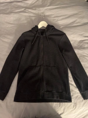Svart zip hoodie med fickor - Säljer en svart hoodie med dragkedja framtill och två stora fickor. Tröjan har huva med dragsnören och är tillverkad i ett mjukt, ribbat material. Perfekt för en avslappnad och stilren look.