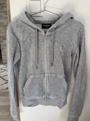 Grå zip hoodie från Juicy Couture - Säljer en grå zip hoodie från Juicy Couture med huva och dragsko. Hoodien har två stora fickor framtill och ett snyggt J-dragkedjesmycke. Mjuk och skön bomullsblandning, perfekt för en chill look.