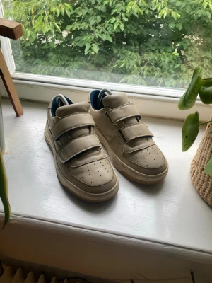 Beige sneakers med kardborre Acne Studios - Snygga beige sneakers från Acne Studios med tre breda kardborreband och mörkblå detaljer. Skorna har perforerad tå, chunky sula och Acne Studios-logga på bandet. Tillverkade i slätt läder med vadderad plös och häl.