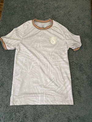 Vit mönstrad t-shirt med randiga detaljer - Säljer en vit t-shirt med diskret geometriskt mönster och randiga detaljer i orange, svart och vitt på krage och ärmslut. T-shirten har korta ärmar och en liten logga på bröstet. Perfekt för en avslappnad och stilren look. Galatasaray fotbollslag