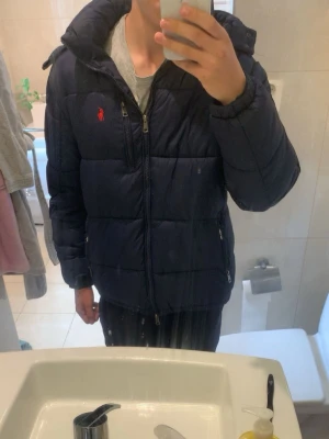 Mörkblå ralph lauren jacka - Snygg mörkblå pufferjacka från Polo Ralph Lauren med röd logga på bröstet. Jackan har dragkedja framtill, två sidofickor med dragkedja och en hög krage med huva. Perfekt för kalla vinterdagar och riktigt skön att bära. Den har en liten deffekt runt vardera ficka men inget som syns