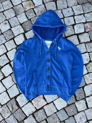 Abercrombie & Fitch cardigan - Snygg blå hoodie med knappar framtill och dragsko i huvan. Har två fickor och ett vitt broderat älgmotiv på bröstet. Tillverkad i mjukt sweatshirtmaterial som passar perfekt till en avslappnad stil. Storlek L enligt etiketten men sitter som S
