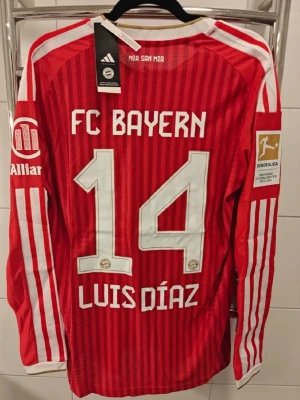 FC Bayern München spelar version matchtröja röd - Säljer en långärmad röd FC Bayern München matchtröja från Adidas med vita detaljer och tryck. Tröjan har klubbmärke, sponsorloggor och nummer 14 med namnet Luis Díaz på ryggen. Perfekt för dig som älskar fotboll och vill stötta Bayern.