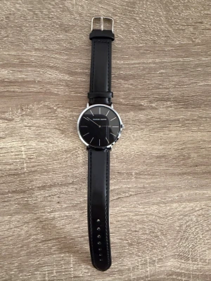 Svart klocka från Hannah Martin - Stilren klocka från Hannah Martin med svart urtavla och silverdetaljer. Klockan har ett runt boett i silverfärg och ett svart, slätt armband i syntetmaterial. Minimalistisk design utan siffror, perfekt för dig som gillar enkel och modern stil.