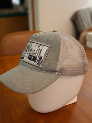 Grå velvet keps med stadsmotiv - Snygg grå keps med mesh på sidorna och baksidan för extra ventilation. Framtill finns en patch med ett svartvitt stadsmotiv som ger kepsen en unik och cool look. Materialet framtill är mjukt och mockaliknande, perfekt för en avslappnad stil
