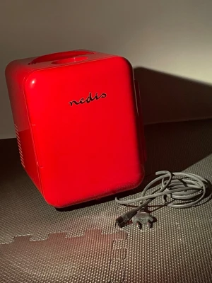 Röd minikyl från Nedis - Snygg röd minikyl från Nedis, perfekt för att förvara hudvårdsprodukter och skönhetsverktyg. Kylen har en kompakt design med rundade hörn, ett bärhandtag på toppen och en hylla inuti för smart förvaring. Tillverkad i plast och metall, med medföljande strömkabel.