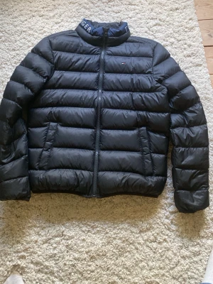 Svart pufferjacka från Tommy Hilfiger - Snygg svart pufferjacka från Tommy Hilfiger med diskret logga på bröstet och märkning på kragen. Jackan har hög krage, dragkedja framtill och två sidofickor. Perfekt för kalla vinterdagar och riktigt stilren look.