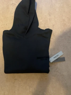 Svart Essentials Fear of God hoodie -  Svart hoodie från Essentials Fear of God helt äkta och är verified med stor logga på ryggen och mindre logga framtill. Klassisk passform med luvan och väldigt varm har även och helt ny med plasticbagen fick den i present men passar inte mig ny pris ligger på 2500kr. (Priset kan diskuteras såklart)