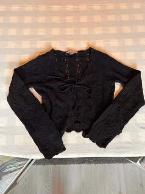 Svart virkad kofta med knytband - Trendig svart virkad kofta med långa ärmar och snyggt knytband framtill. Koftan har ett vågigt avslut nertill och ett genombrutet mönster som ger en cool vintagekänsla. Perfekt att slänga över ett linne eller topp för en extra touch.