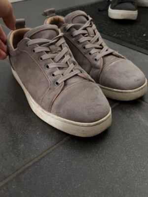 Gråa loubs  - Snygga gråa sneakers i mocka från Christian Louboutin med klassisk röd sula och vita kanter. Skorna har rund tå, matchande gråa skosnören och diskreta sömdetaljer. Perfekta för dig som gillar stilrena och exklusiva sneakers. Kommer med kvitto
