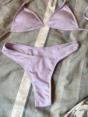 Ljuslila ribbad bikini - Säljer en ljuslila bikini med ribbad struktur. Överdelen har trekantskupor med tunna band att knyta i nacken och ryggen. Underdelen är enkel och högt skuren. Perfekt för sommardagar på stranden eller vid poolen.