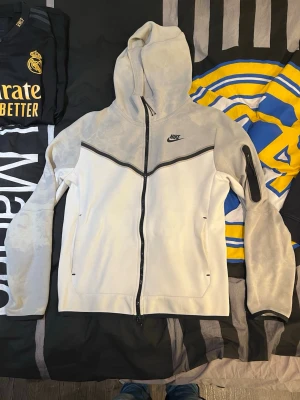 Nike tech fleece - Nike tech hoodie i vitt och grått med svarta detaljer och dragkedja framtill. Har en huva, två sidofickor och en extra ficka med dragkedja på ena ärmen. Snygg design med svart linje över bröstet och Nike-logga på vänster sida. Perfekt för en sportig och avslappnad stil. ( fläckar som inte går bort i tvätten ) därav är den så billig