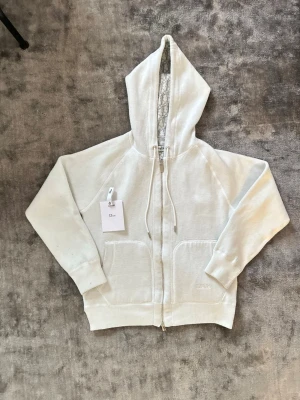 Vit hoodie med dragkedja Di0r - Säljer en vit hoodie Di0r med dragkedja framtill och två stora fickor. Huvan har ett snyggt mönstrat foder med Di0r-logga och justerbara snören. Ribbstickade muddar vid ärmslut och nederkant. Perfekt för dig som gillar exklusiv streetwear.